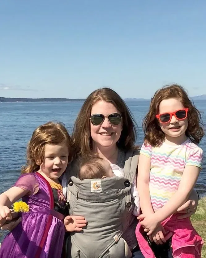 Megan Taylor, a CRNA, casually dressed at lake with her two toddler daughters next to her and an infant in a sling.