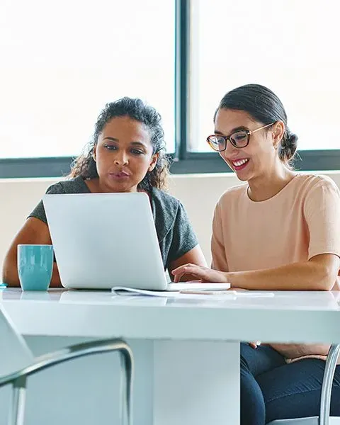 Multiethnic medical residents, both women, look at a laptop screen smiling because of articles on PS&D's GME Resource Lounge.