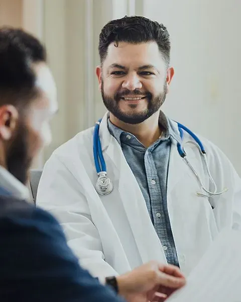 A physician, in a white coat and stethoscope, talking with a PS&D Recruiter about Locum Tenens options, both are Hispanic.