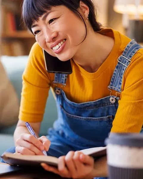 An Asian woman physician on the phone with a friend, celebrating paying off loans, wearing a yellow top and denim overalls.