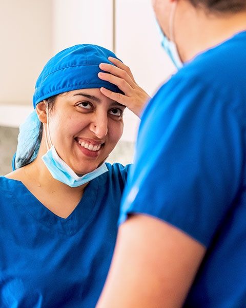 A surgeon, a Persian woman in blue scrubs and face mask, smiling with her colleague, a man, wearing the same.