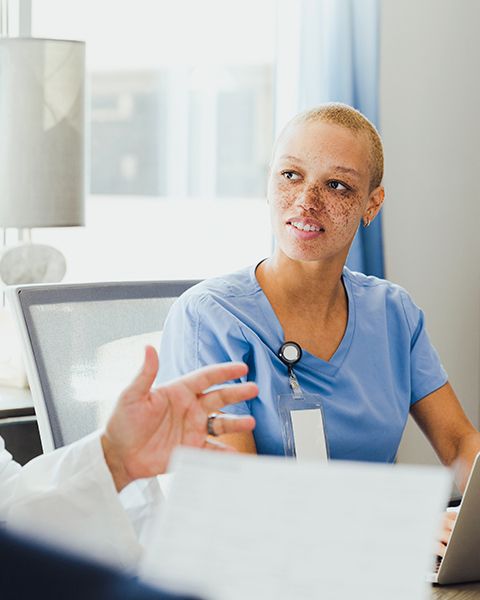 A physician, a Black woman, in blue scrubs have a conversation with another physician.