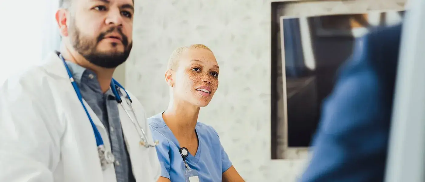 A physician, a Hispanic man, in a white coat and stethoscope next to a Surgeon in blue scrubs, a Black woman with freckles.