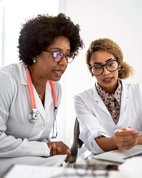 Two physicians, both Black women in white coats, the older woman is consulting on a patient case with the younger woman.