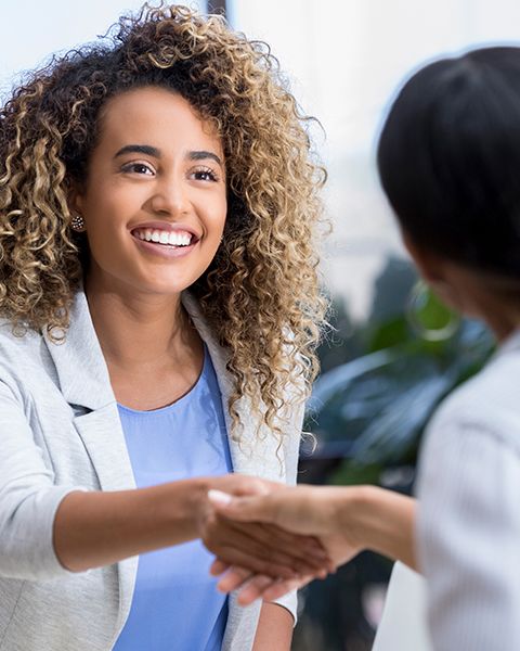 A physician, a Black woman, in natural curls and a professional white suit shaking hands with her interviewer, a Black woman.