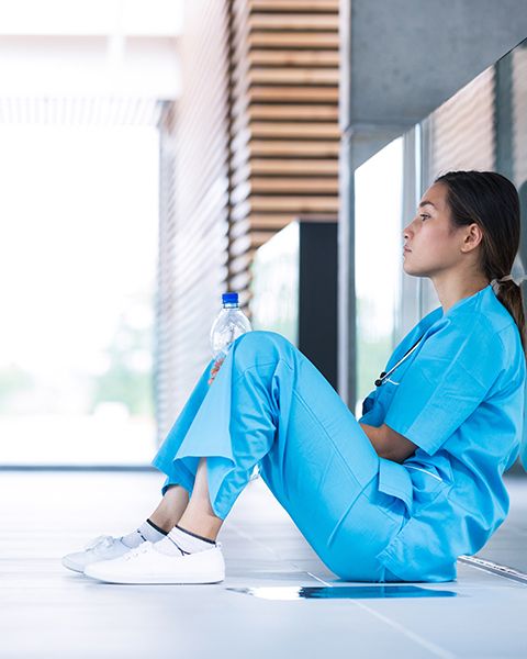 A physician, a woman of Arab descent, sits on the floor in blue scrubs, feeling self-doubt and imposter syndrome.